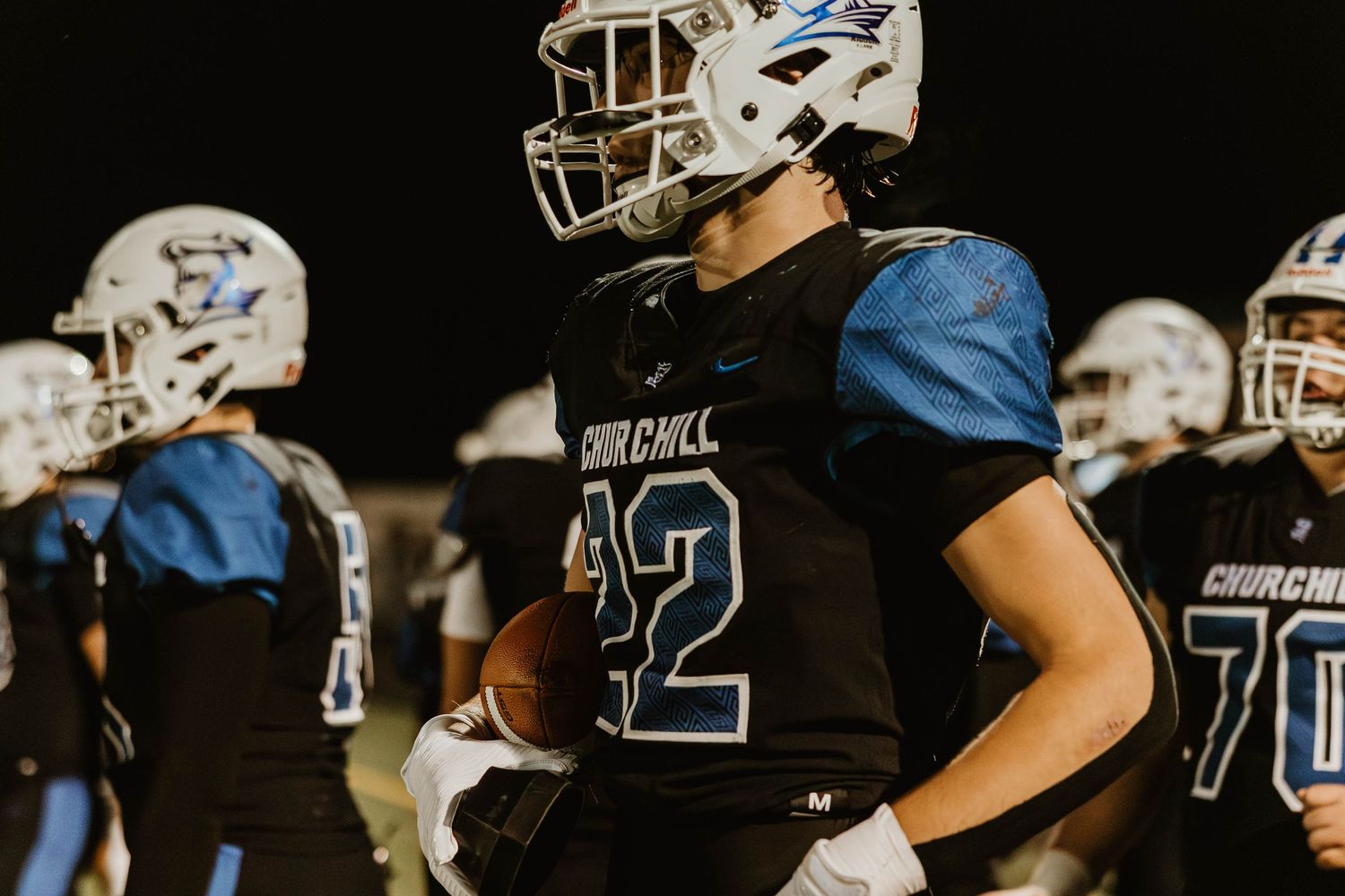 Football players in black and blue uniforms line up on the field during a nighttime game.