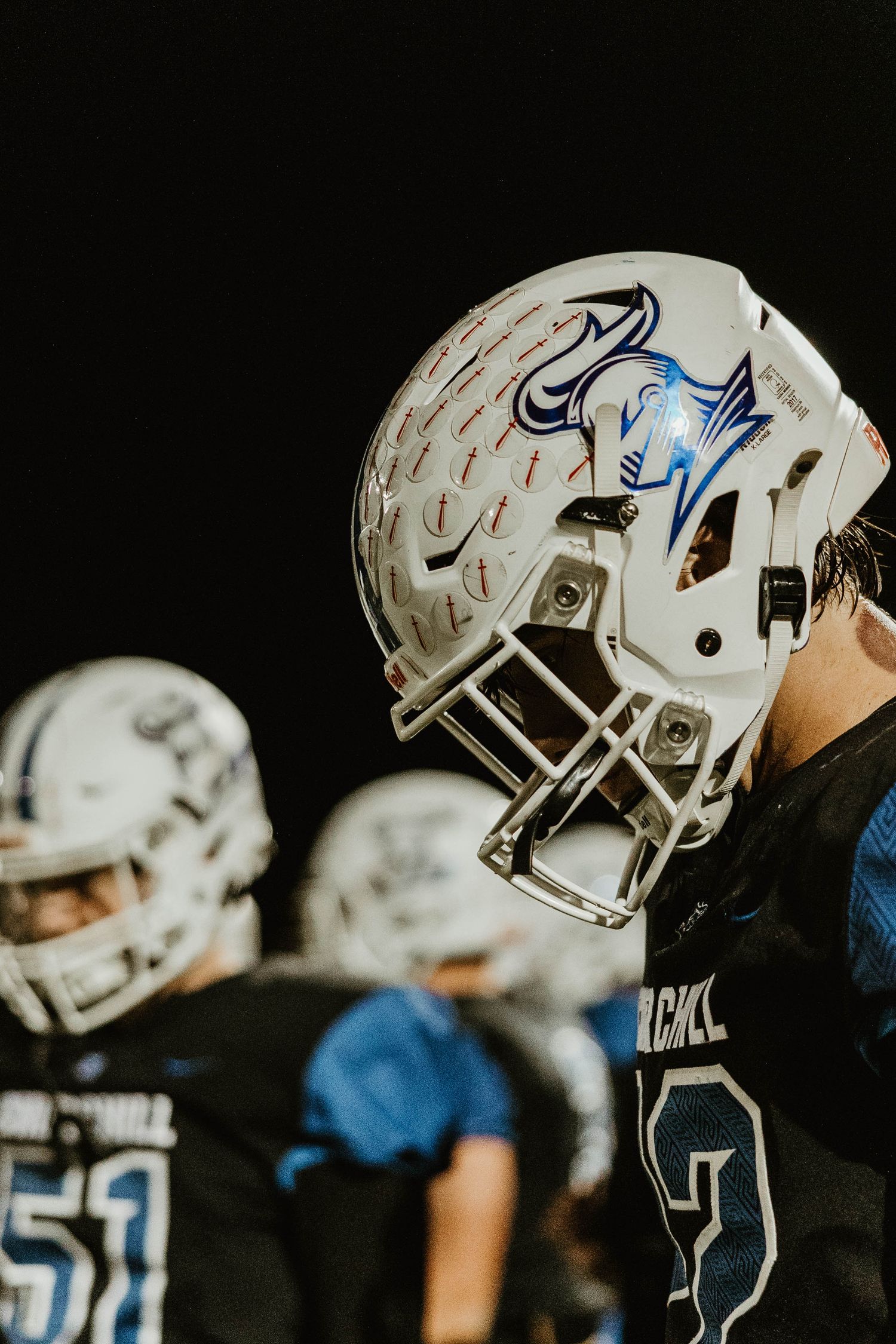 Row of white football helmets with blue accents lined up on the Churchill team sideline.