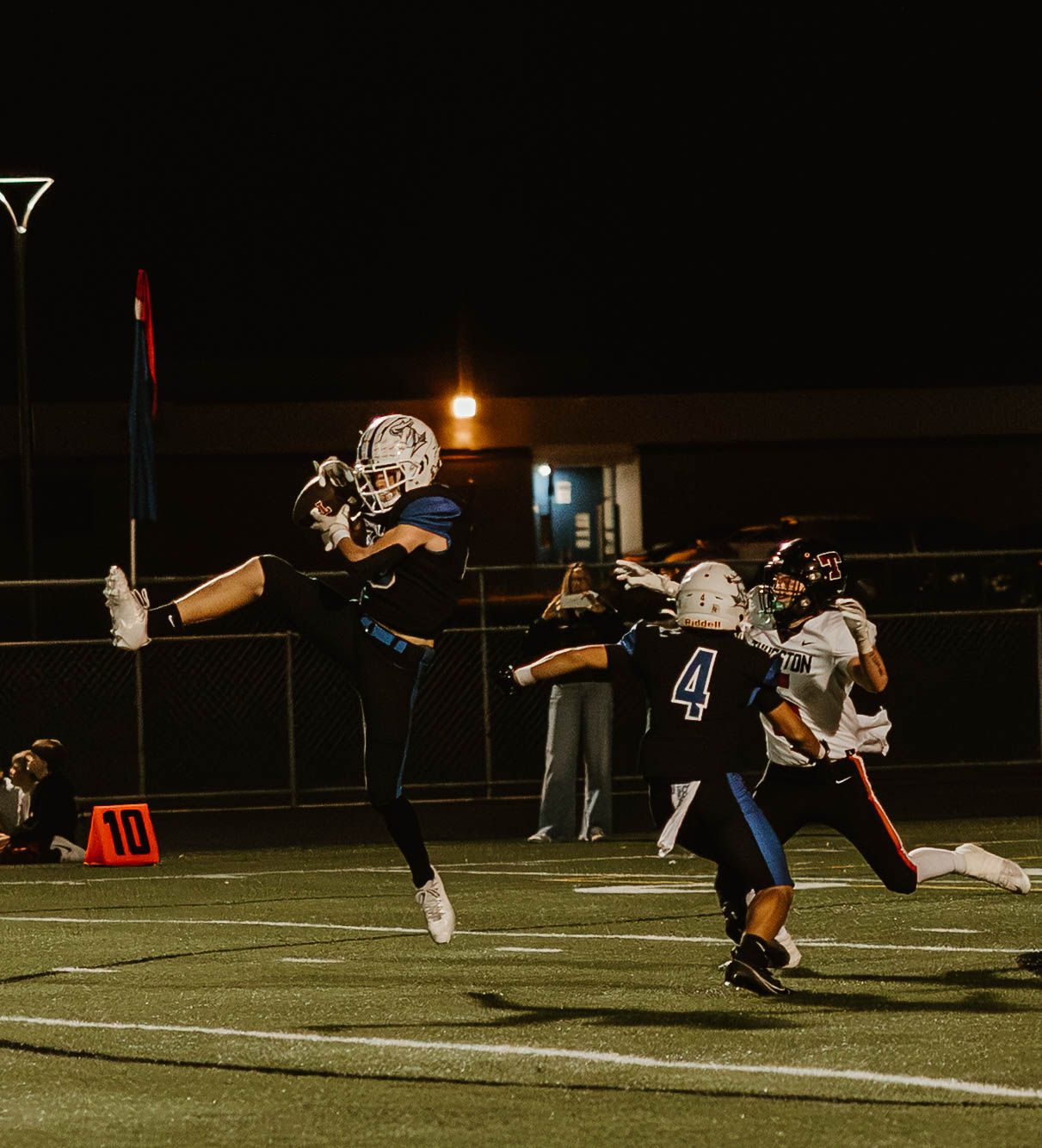 High school football players leap and compete for the ball during a nighttime game under stadium lights.