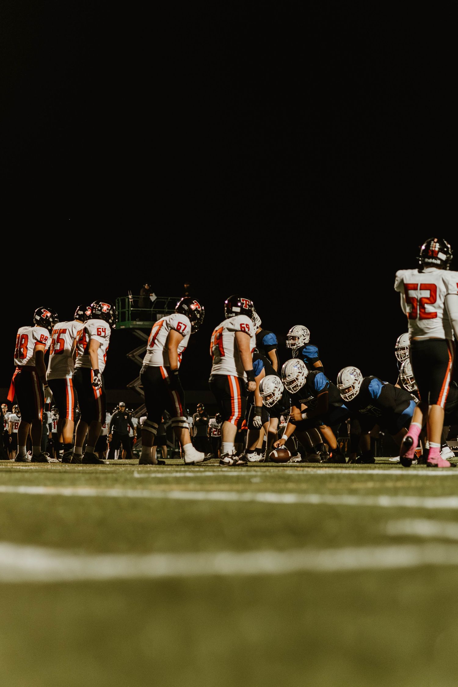 Football players line up at the line of scrimmage during a nighttime game under stadium lights.