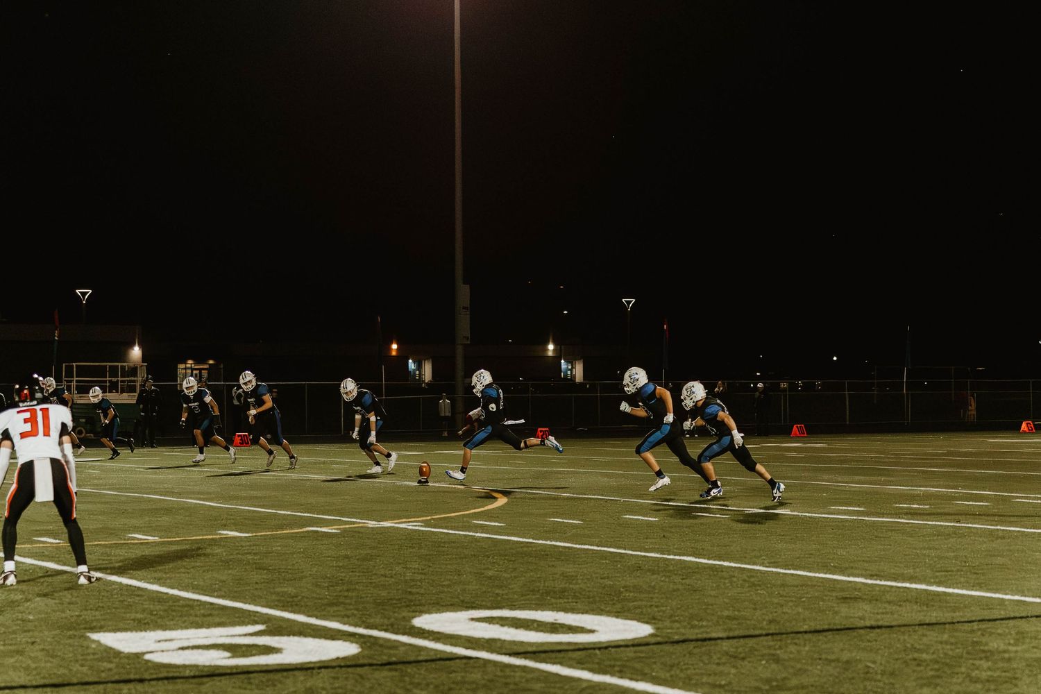 Football field illuminated by bright stadium lights at night with players scattered across the turf.