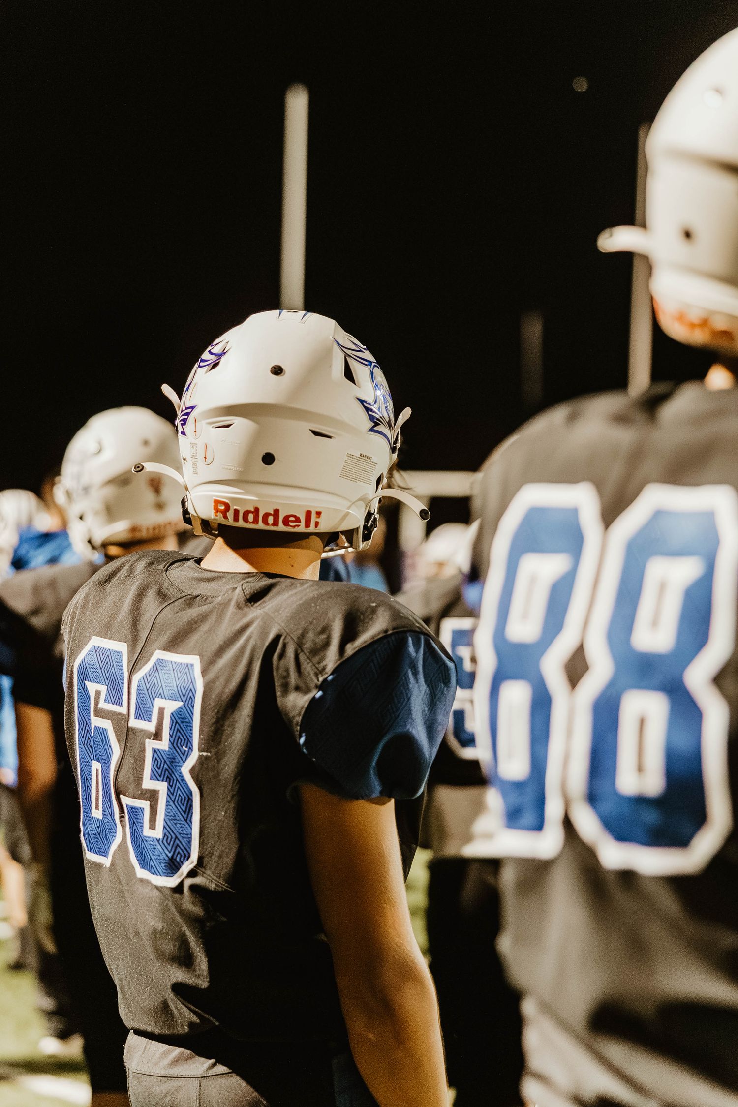 Football players in gray jerseys line up on the sideline showing jersey numbers in blue and white during a night game.