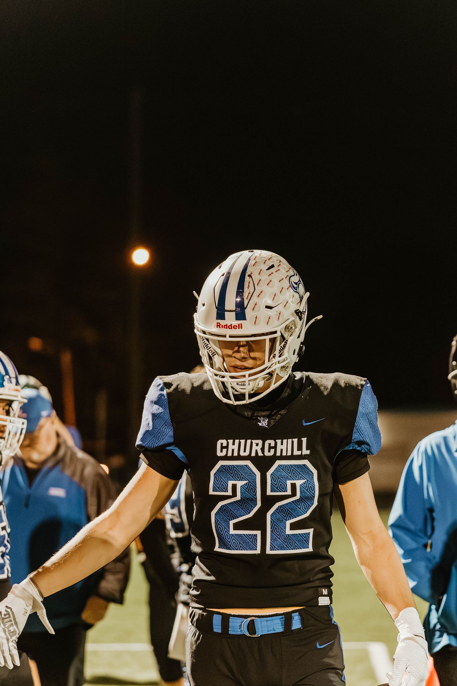 Football players in black and light blue uniforms line up during a nighttime game at Churchill stadium.