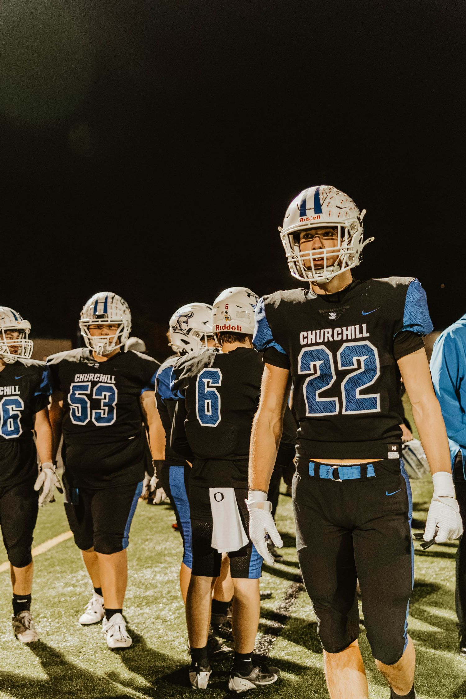 Churchill football team players in black and blue uniforms stand together on the field at night.