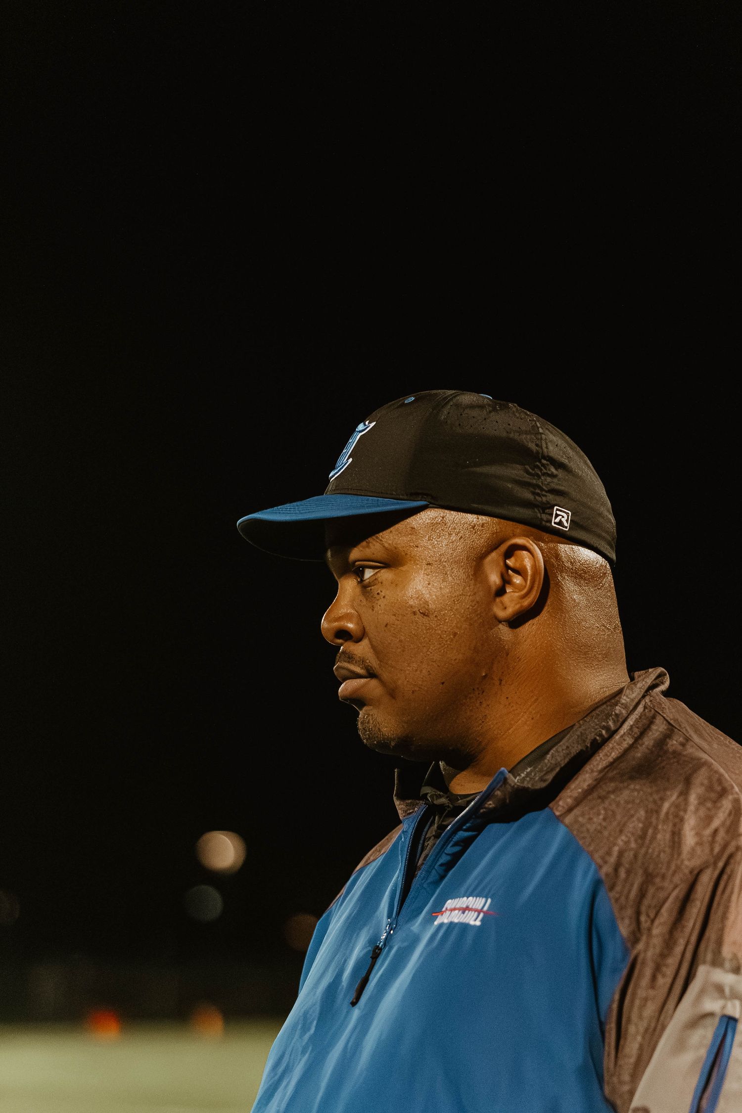 Football coach in a blue jacket and brown cap observes from the sidelines during a nighttime game.
