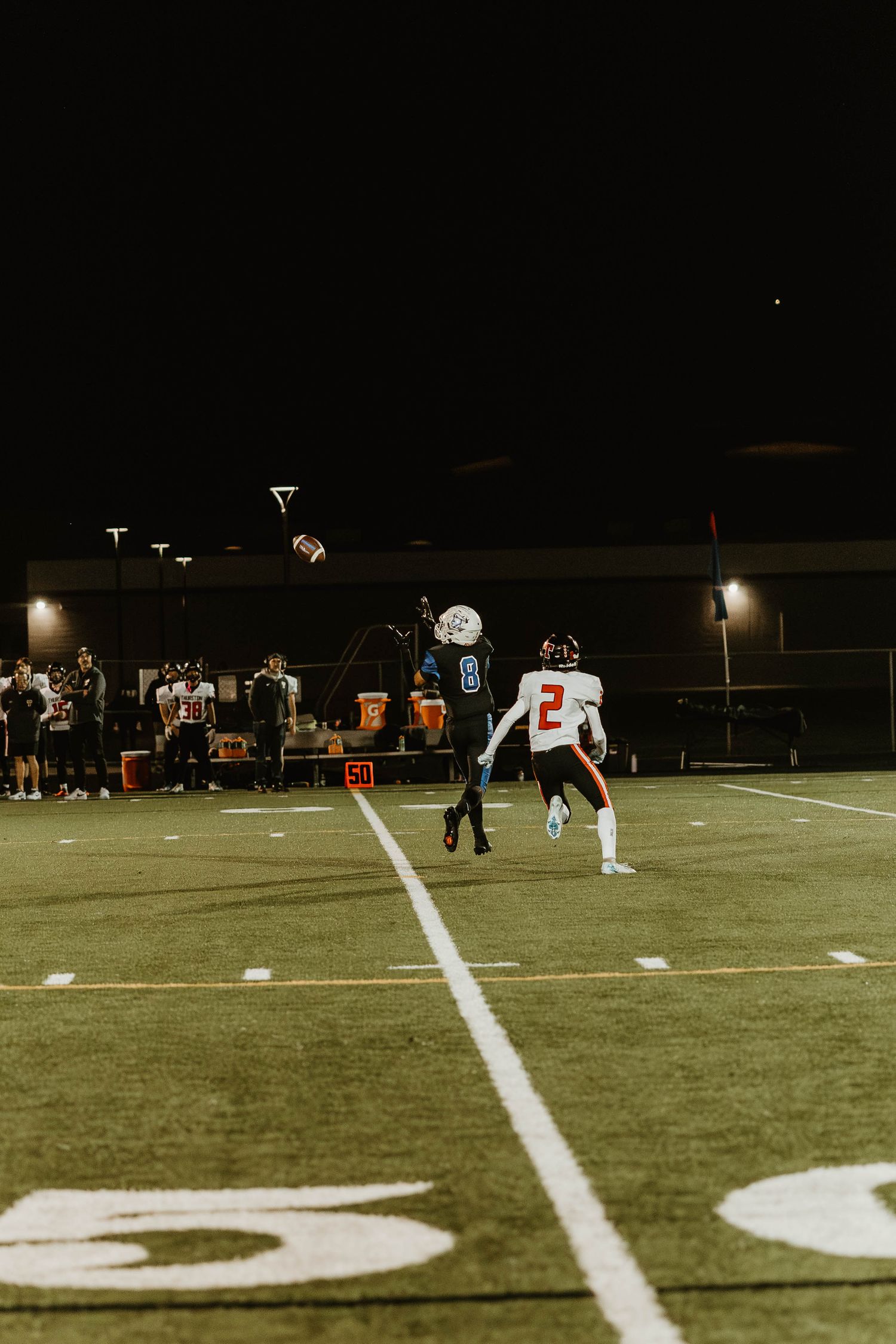 Night football game showing yard line markers and players on the illuminated field.