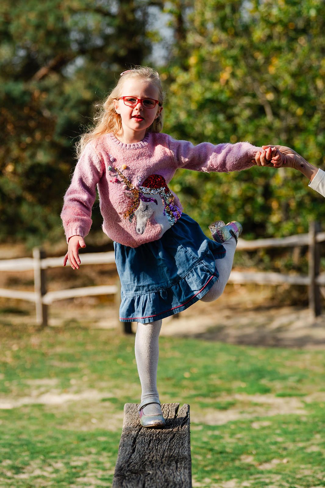 A child in a pink sweater and denim dress practices balancing on a tree stump in an outdoor setting.