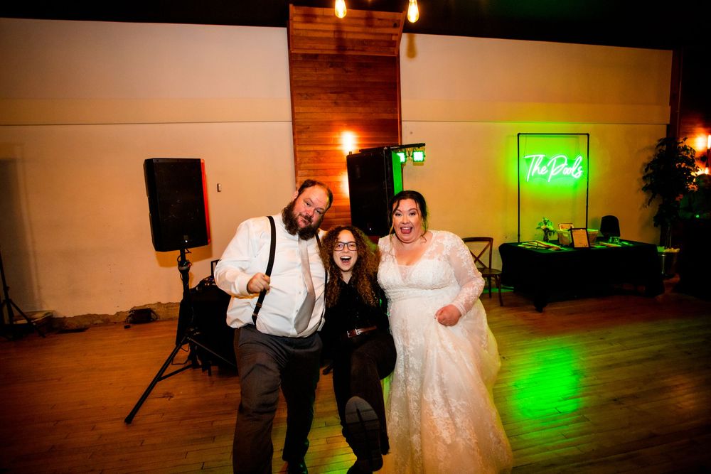 A group of wedding guests dance together on a dance floor illuminated by green lighting at an evening reception.