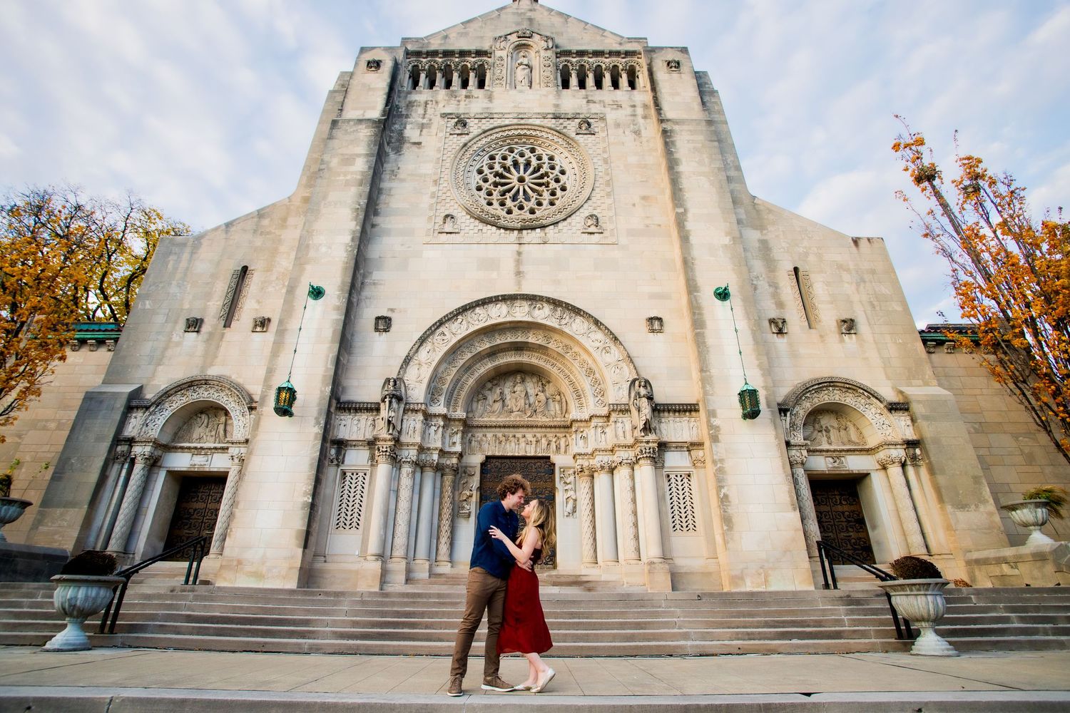 Couple embracing in front of a grand stone cathedral with ornate arches and rose window during fall season.