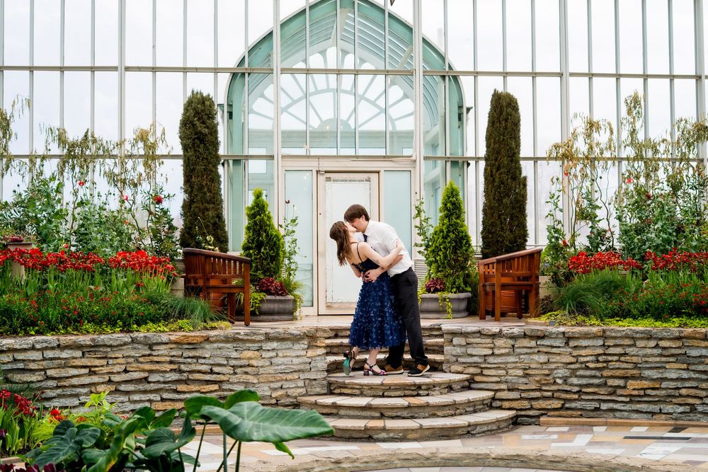 Couple sharing a romantic kiss on stone steps in front of a beautiful greenhouse with red flowers.