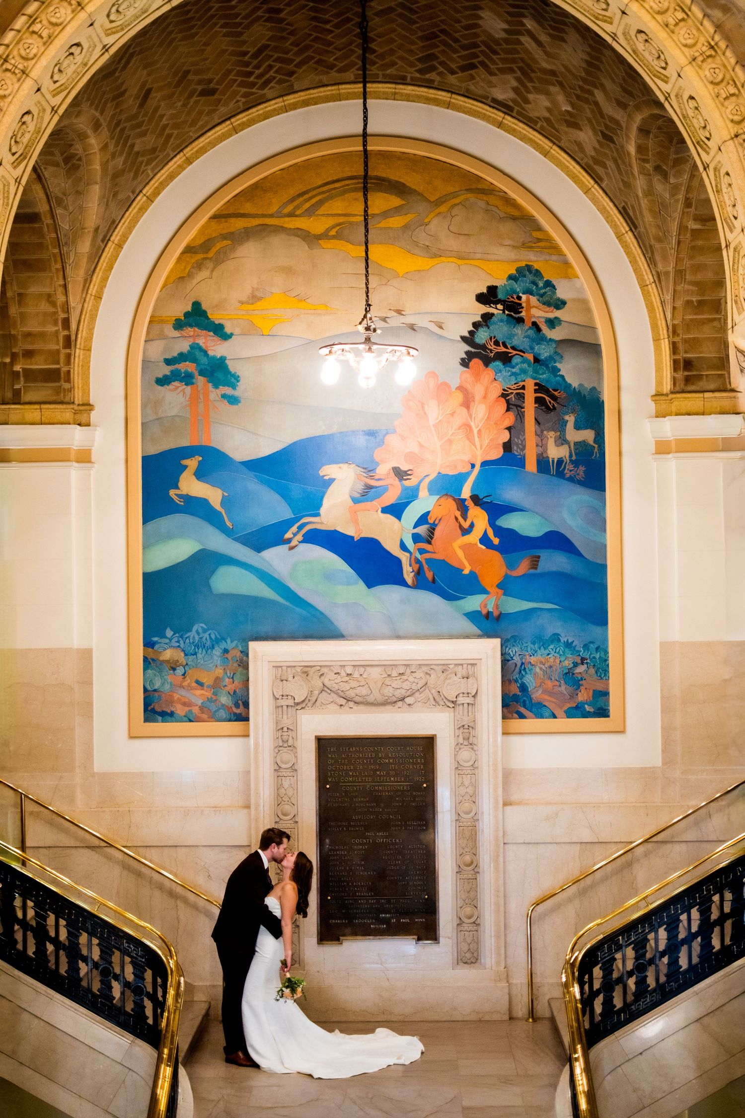 Elegant couple embraces in front of ornate mural with horses and decorative archway inside historic building.