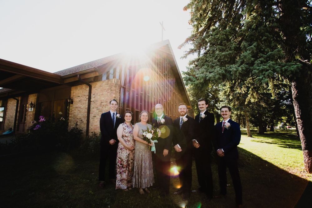 Outdoor wedding ceremony under golden sunlight with guests seated on white chairs facing a rustic brick building.