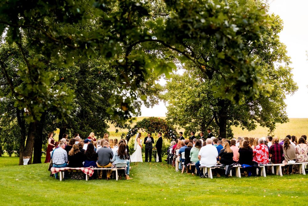 Outdoor wedding ceremony under large oak trees with guests seated on benches on a green lawn.