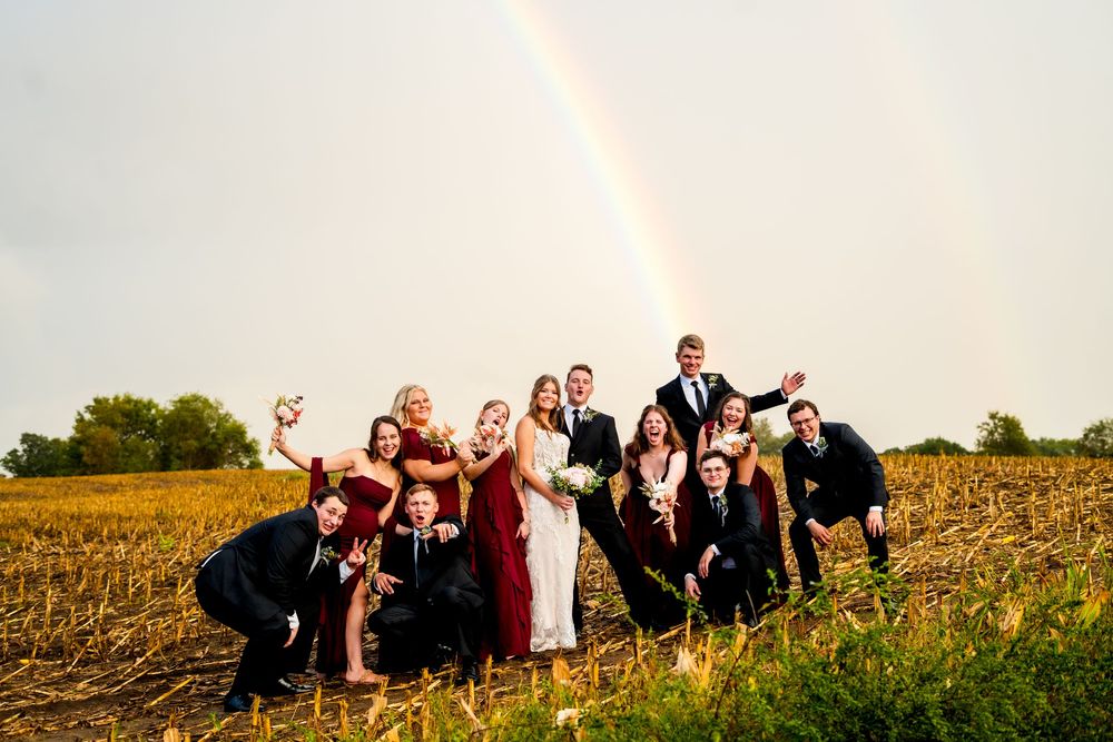 Wedding party poses playfully in a golden field at sunset with a vibrant rainbow arching across the cloudy sky.