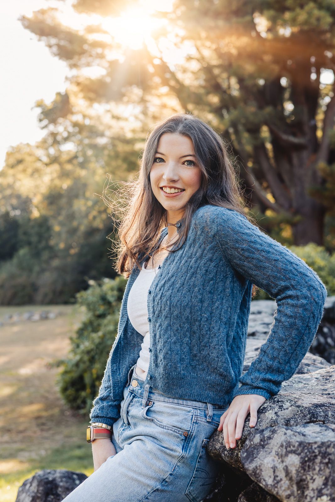 A person wearing a blue knit sweater and jeans poses on rocks during golden hour in a natural outdoor setting.