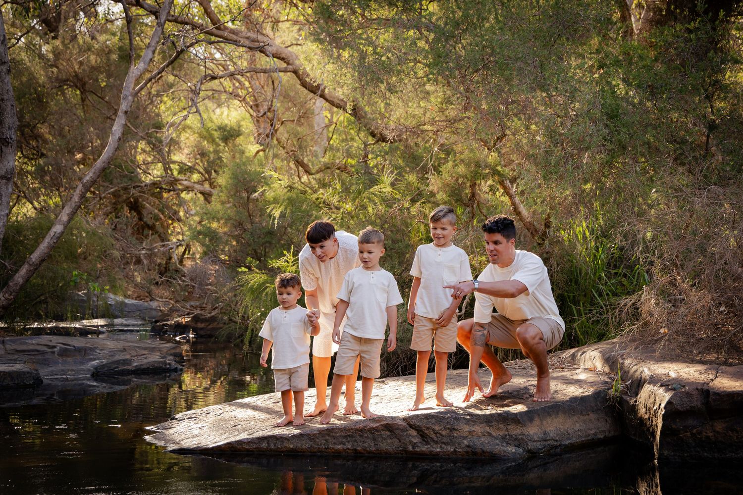Family in white outfits sits together on rocks near a stream during an outdoor photo session at sunset.