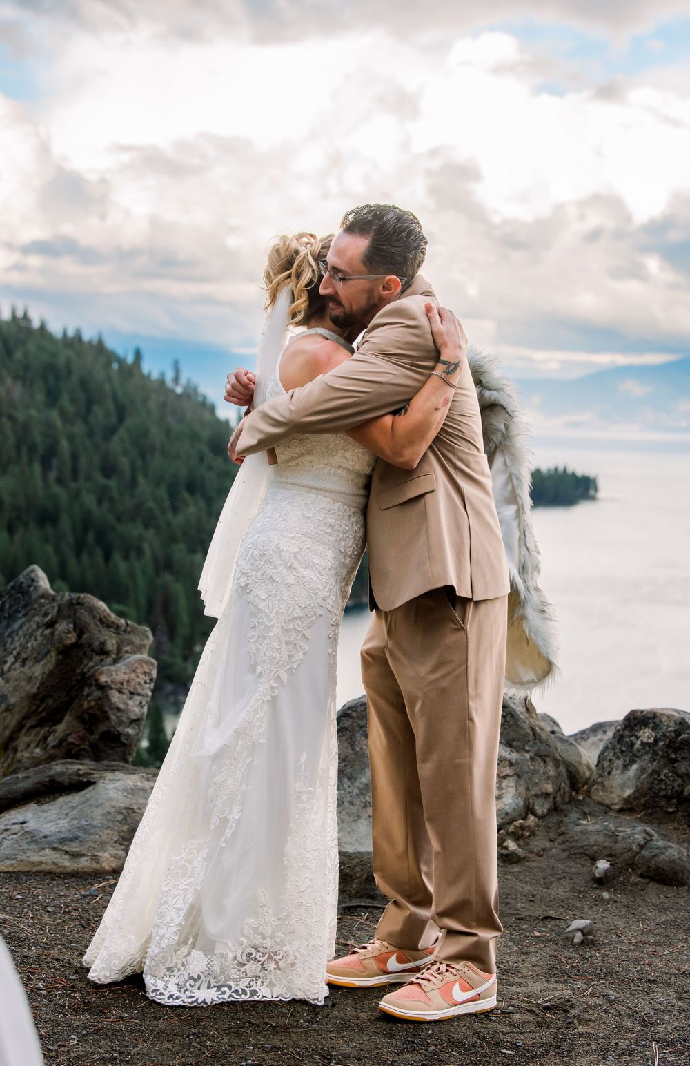Newlyweds embrace on rocky coastal cliffs with evergreen forest and cloudy sky in background.  Lake Tahoe, CA.
