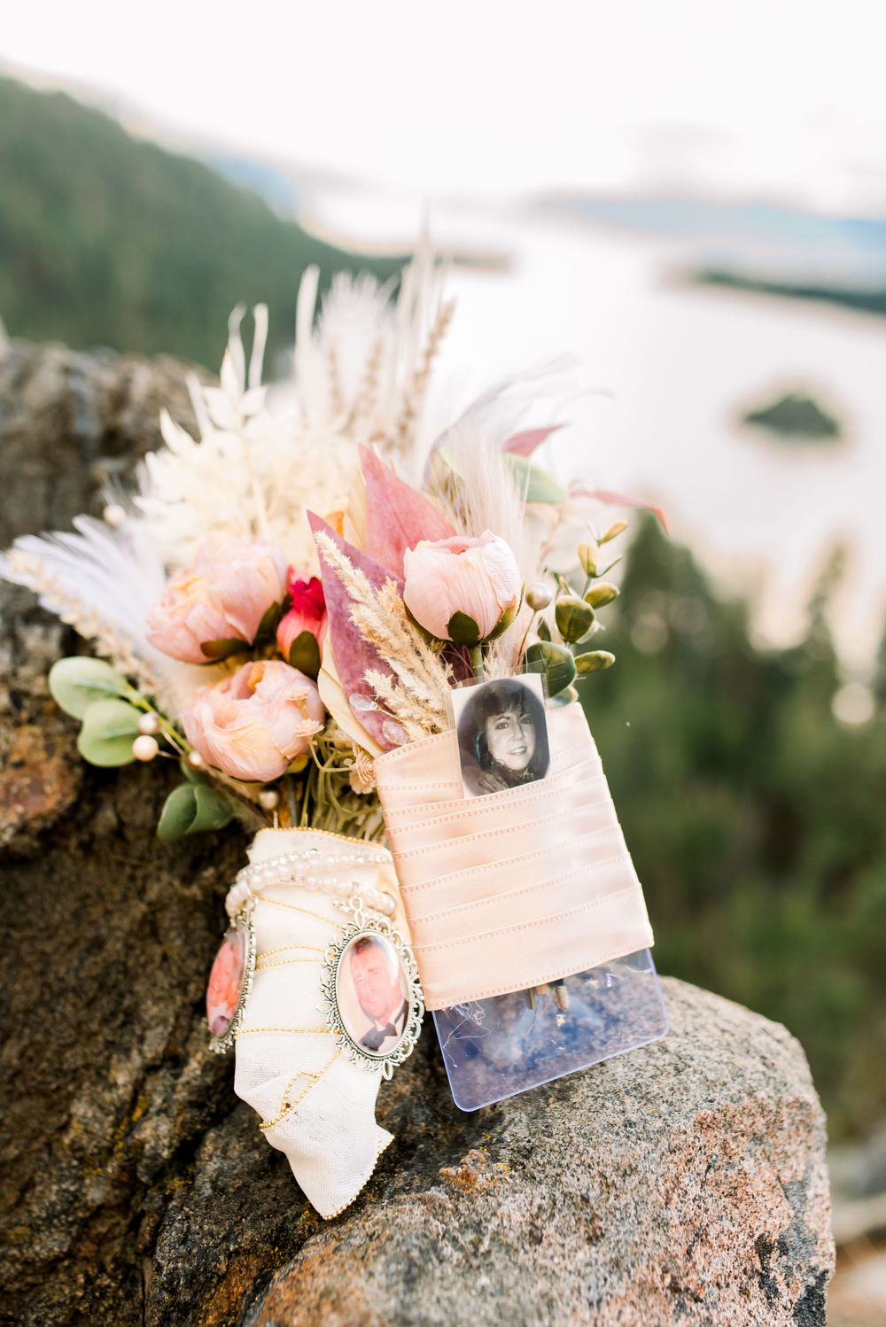 Wedding bouquet with pink roses and dried flowers alongside crystal and agate stones resting on a rocky surface outdoors.