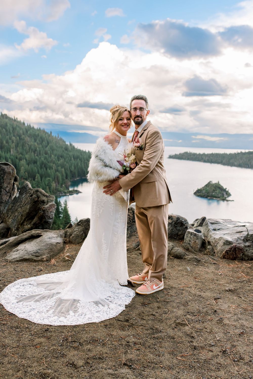 A couple in wedding attire embrace on a cliff overlooking a scenic bay with evergreen forests and islands in the background.