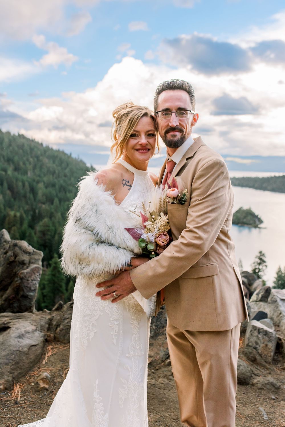 A wedding couple poses on a scenic mountain overlook with a lake and forest backdrop on their special day.