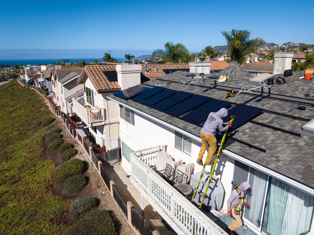 Workers install solar panels on the roof of a house in Pismo Beach with the ocean in the background.