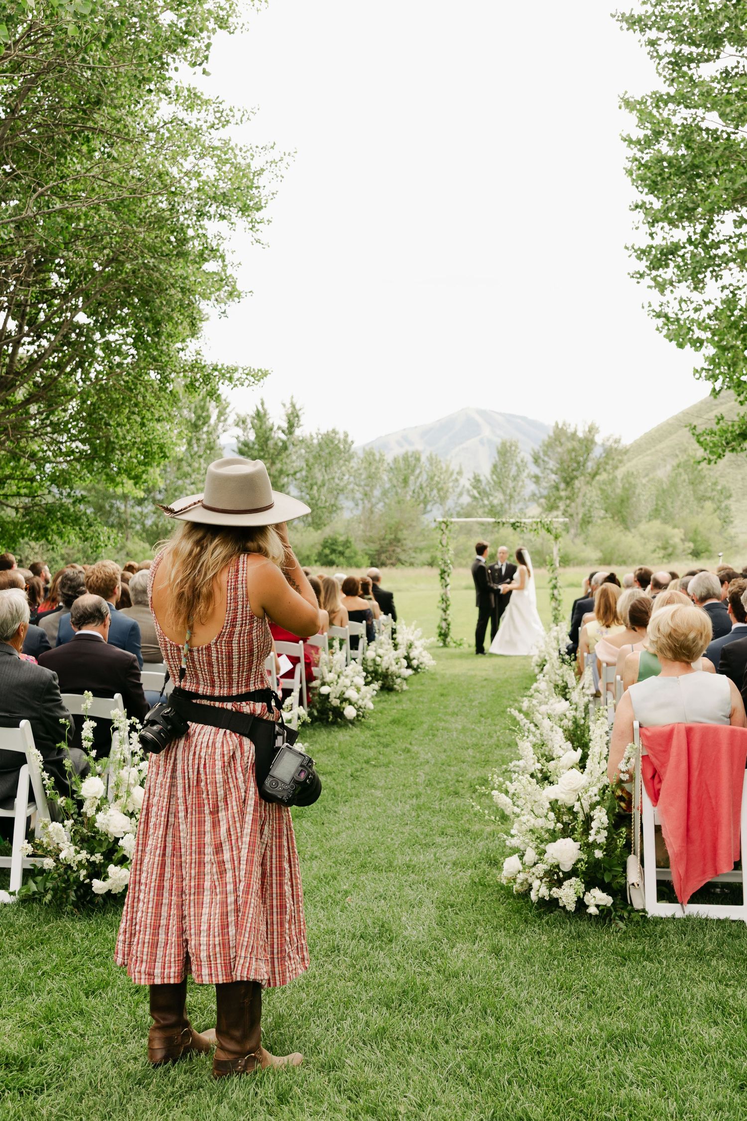 A photographer in a striped dress and hat captures an outdoor wedding ceremony with mountain views and white floral decorations.