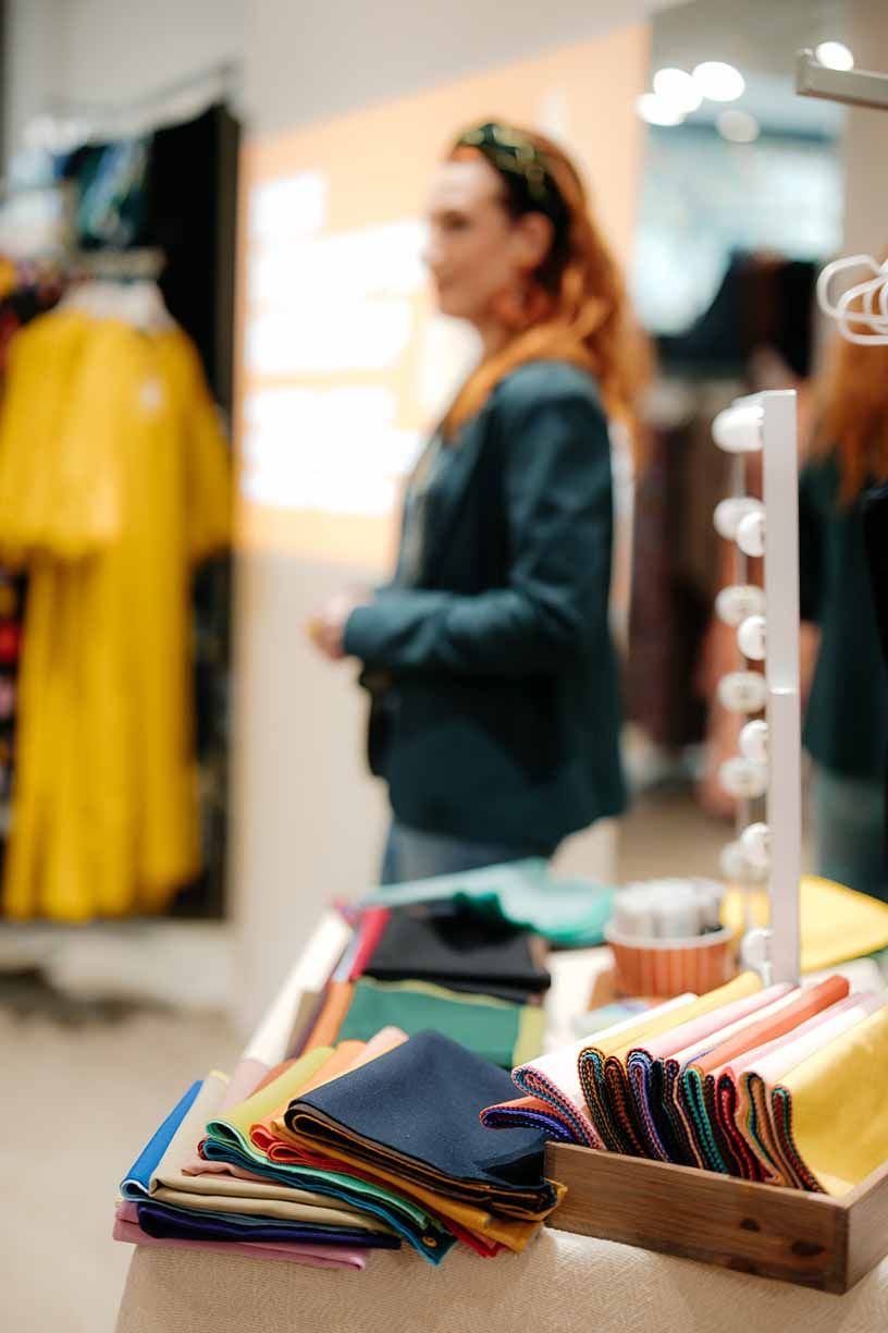 Colorful fabric swatches and samples displayed on retail counter with clothing racks and string lights in background.