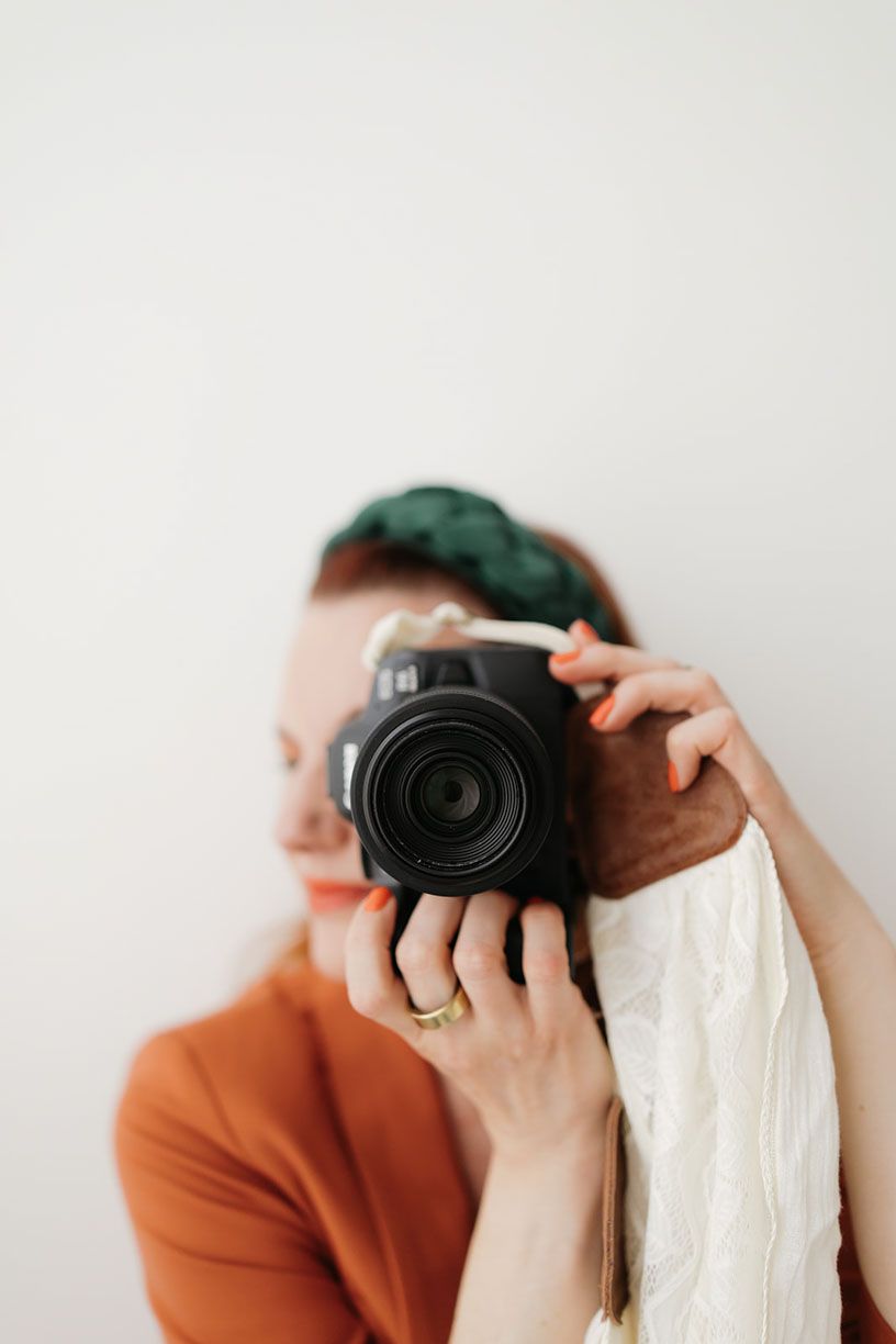Photographer in orange sweater holds camera against white background in minimalist composition series.