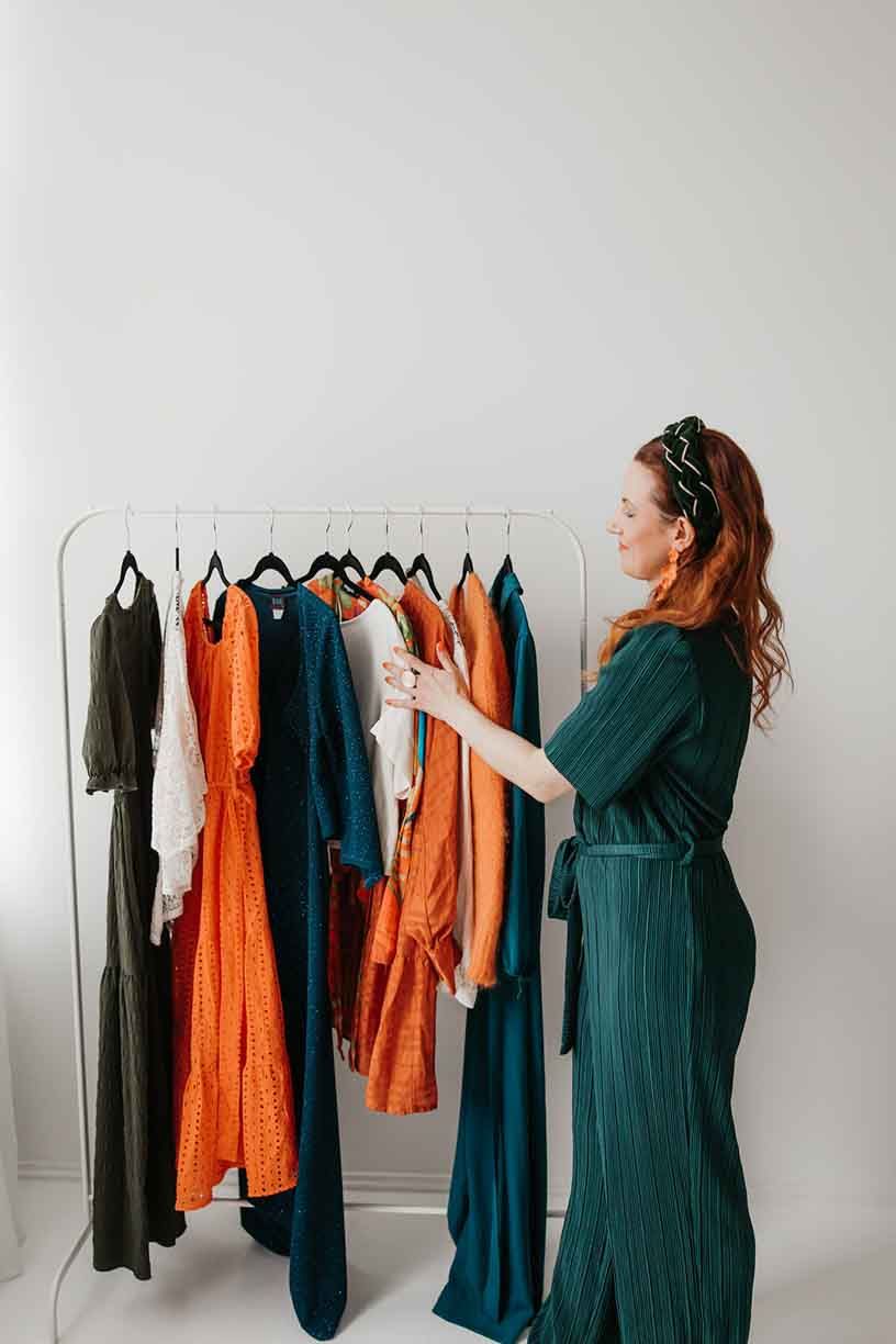 Woman in dark green dress organizing a clothing rack with orange and teal garments against white wall.