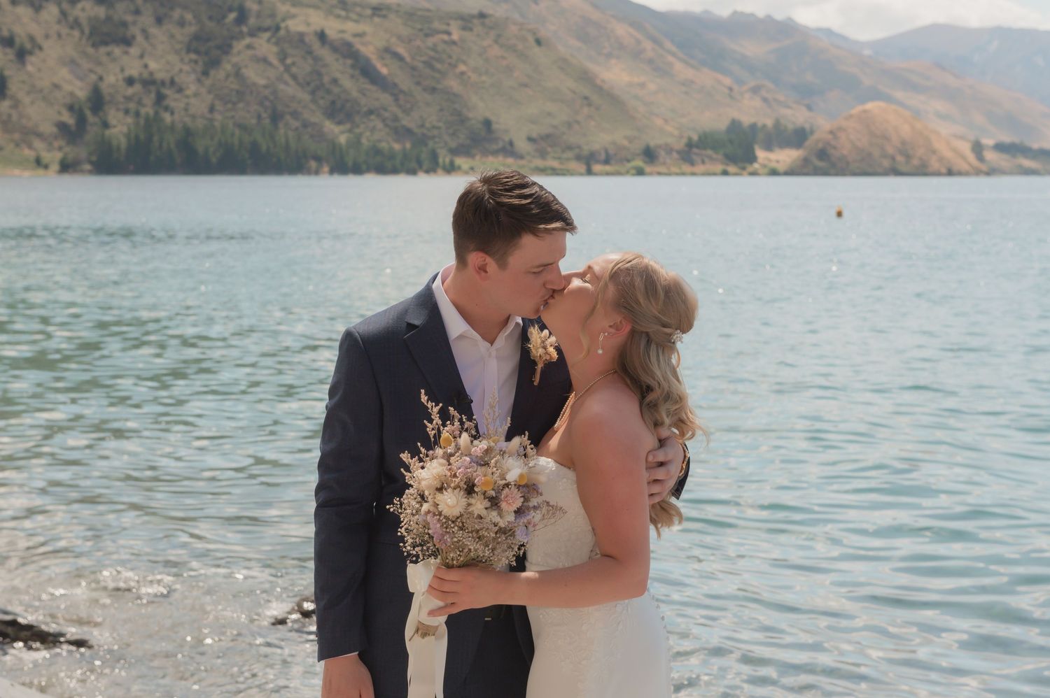 A couple shares romantic moments along a scenic mountain lake shoreline during their wedding photoshoot.