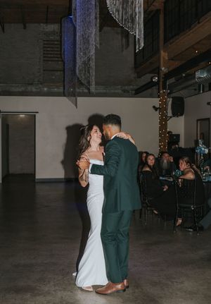 A couple shares their first dance at a dimly lit indoor wedding reception with string lights overhead.