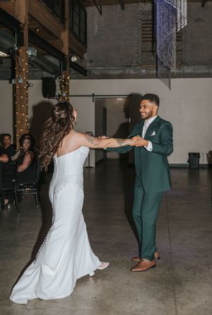 A couple performs a romantic dance sequence in formal wedding attire on a concrete floor at an indoor venue.