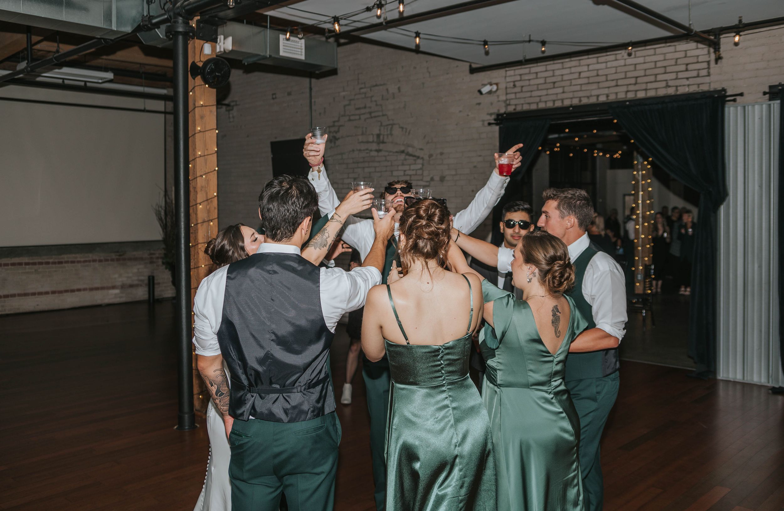 Wedding party dances together on a dark wooden dance floor in an industrial-style venue with exposed brick walls.