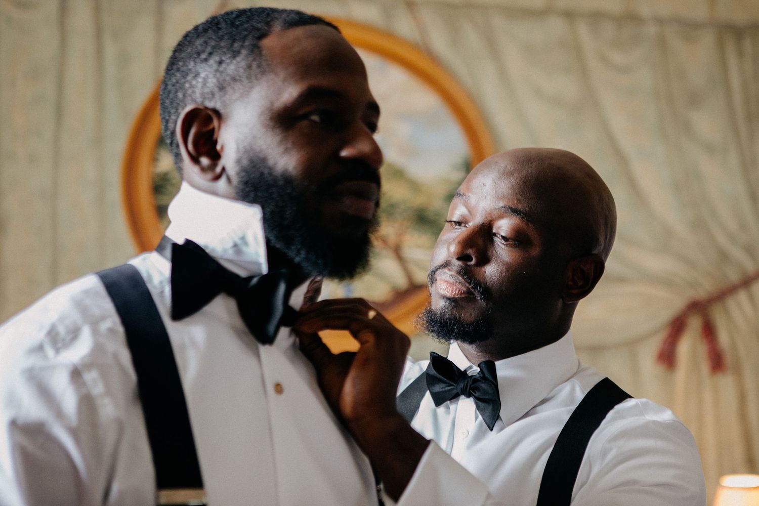Two groomsmen in white shirts and suspenders adjust each other's bow ties before a wedding.