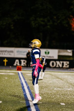 Football player wearing navy and gold uniform kneels on field with pink accessories during night game.