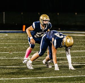Football players in navy and gold uniforms crouch in formation on the field at night.