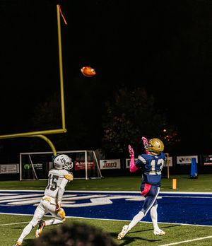 Football player leaps to catch pass in endzone while defender watches during night game.