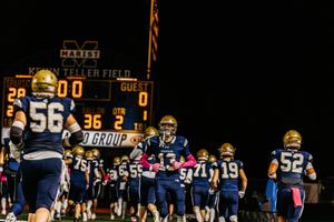 Football team lines up under stadium lights with scoreboard visible in background.