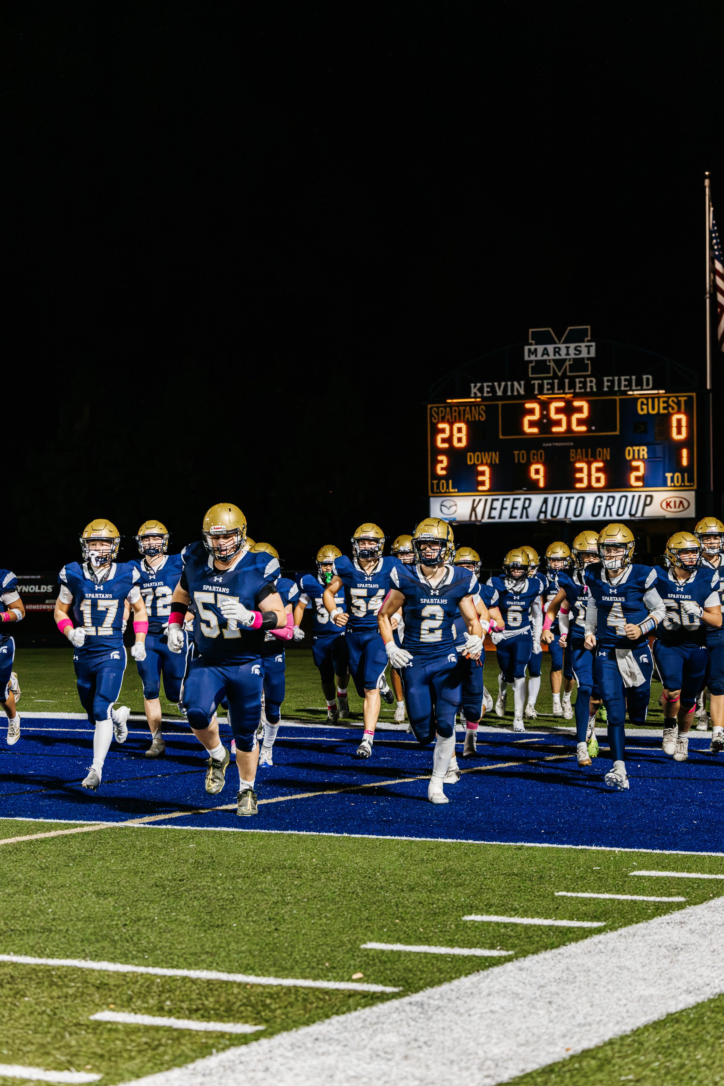 Football team lines up on blue turf field with scoreboard showing in background during nighttime game.