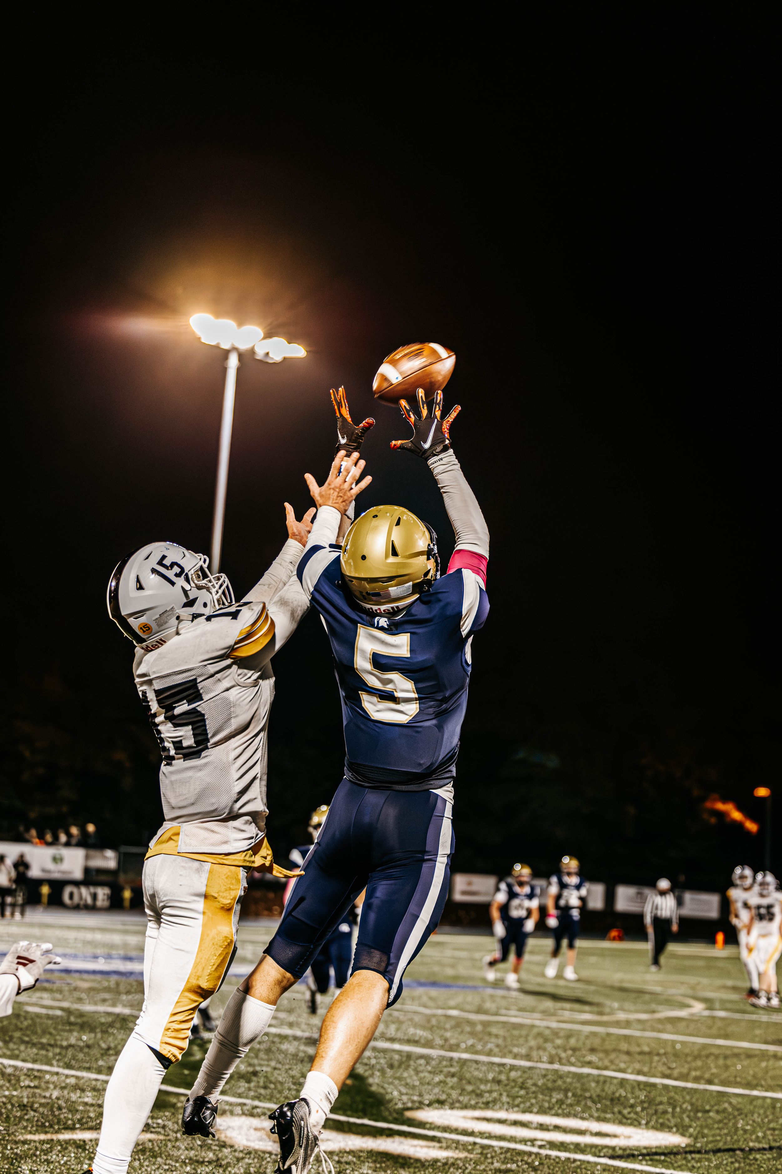 Two football players jump for the ball during nighttime game on illuminated field.