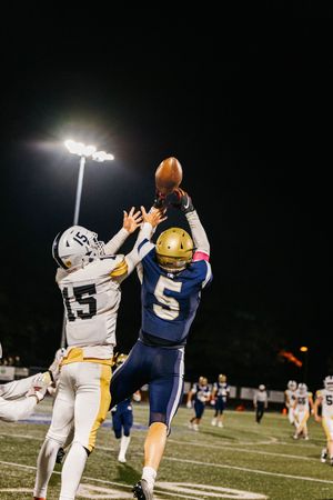 Two football players leap for the ball during a nighttime game under stadium lights.