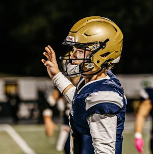 Football player in gold helmet and navy jersey throws pass during nighttime game under stadium lights.