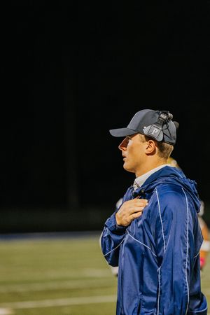 Coaches in blue jackets watch intently from sideline during nighttime football game.