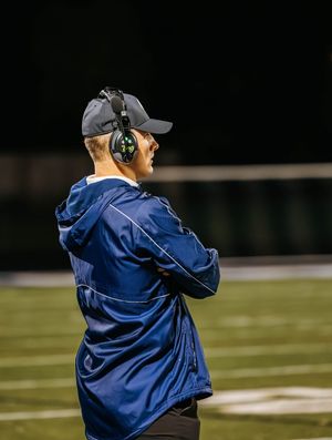 Coach in navy jacket and cap with headset observes from sideline during nighttime game.