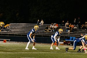Football players line up in formation on a dark field during a nighttime game with spectators in the bleachers.