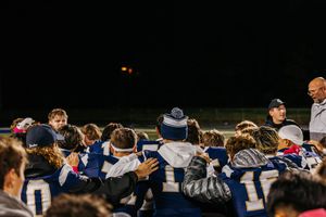 Team huddle of football players in blue jerseys gather with coaches at night.