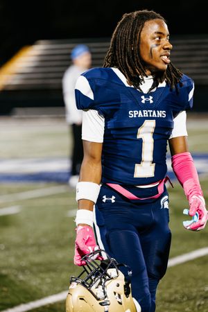 Football player in navy Spartans uniform with pink gloves stands on the field during a game.