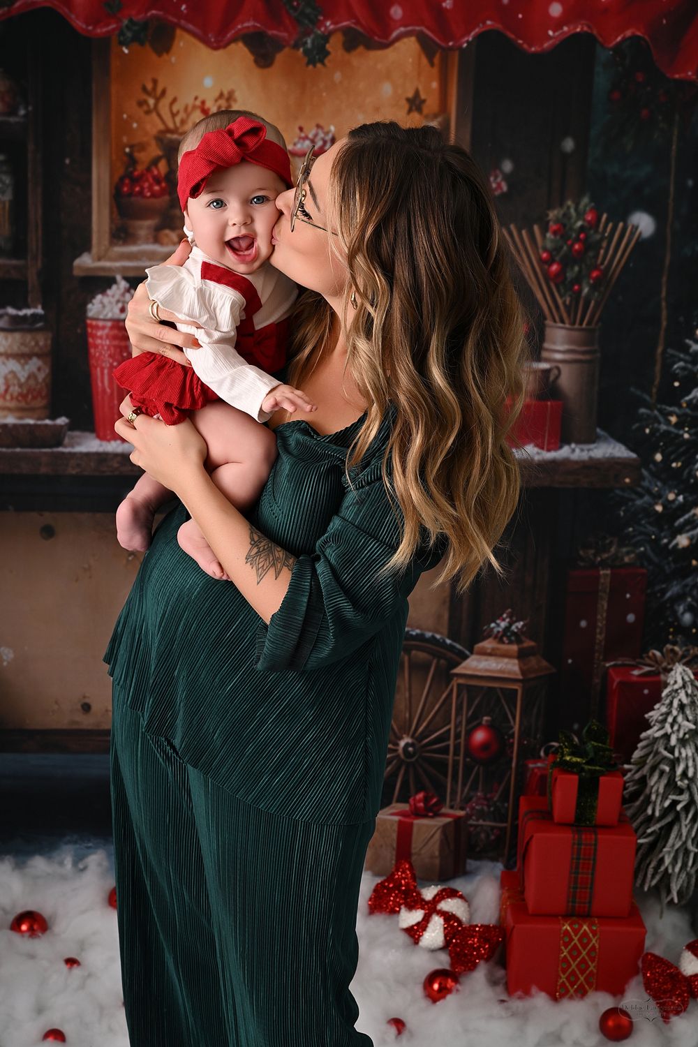 Mother and baby share tender holiday moments in front of a festive Christmas backdrop with red ornaments and presents.