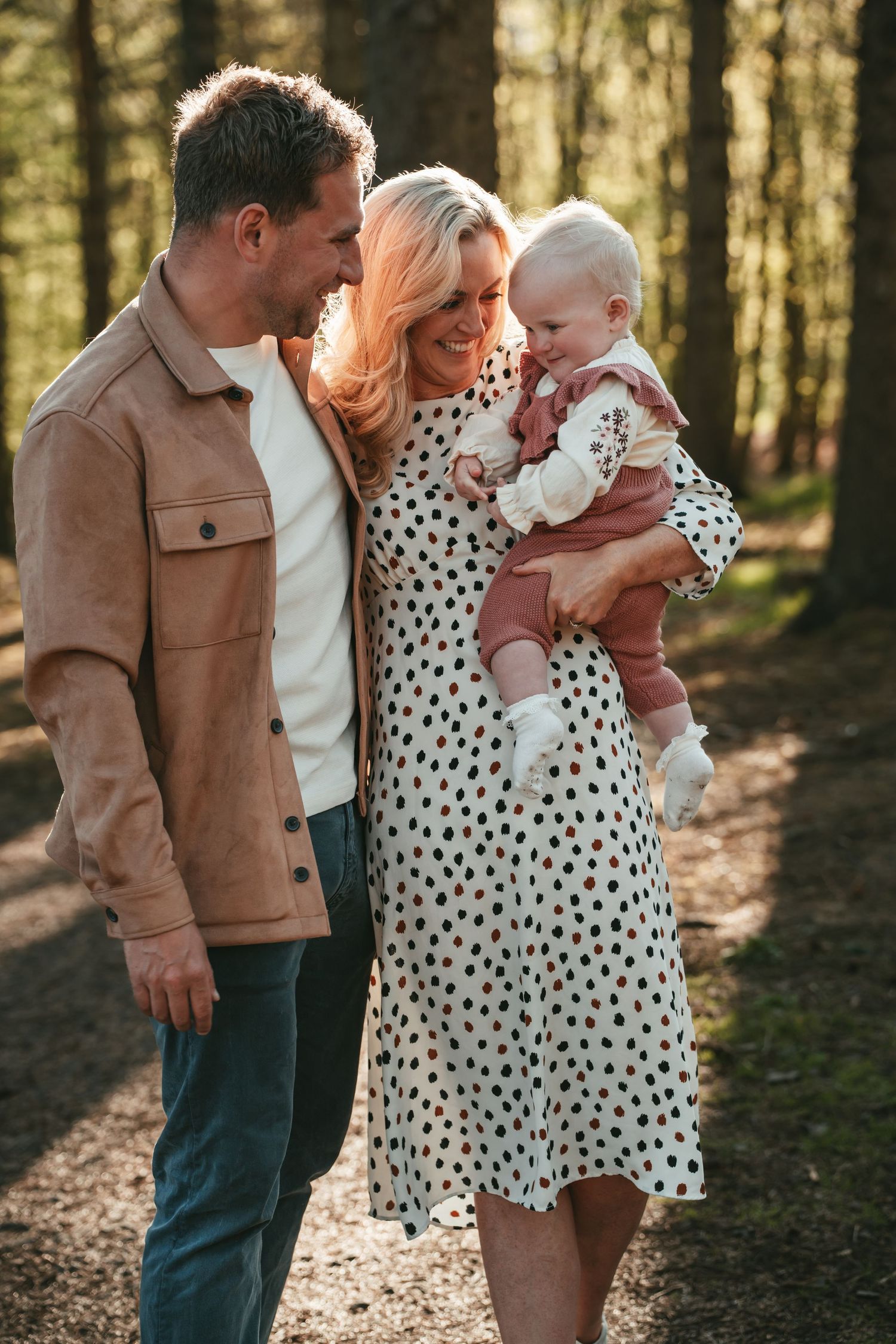 A family shares tender moments together in a sunlit forest setting, wearing casual clothing and polka dot dress.