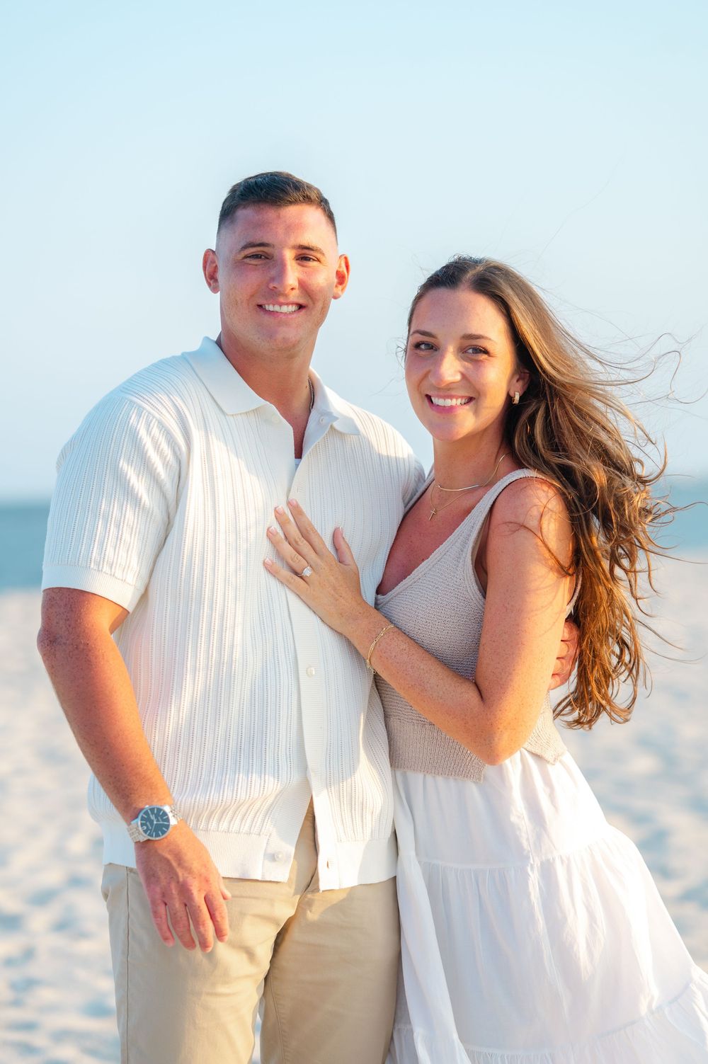 A couple in coordinating white outfits poses together on a sunny beach after getting engaged in Long Island.